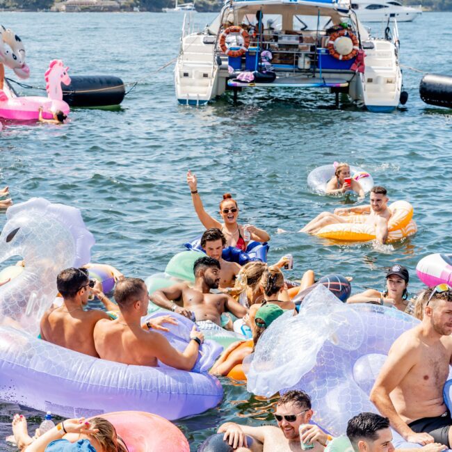 A group of people enjoying a sunny day on inflatable floats in the water near a boat. The floats are colorful, and some are shaped like animals. In the background, a catamaran is moored, and a few more people are relaxing on it.