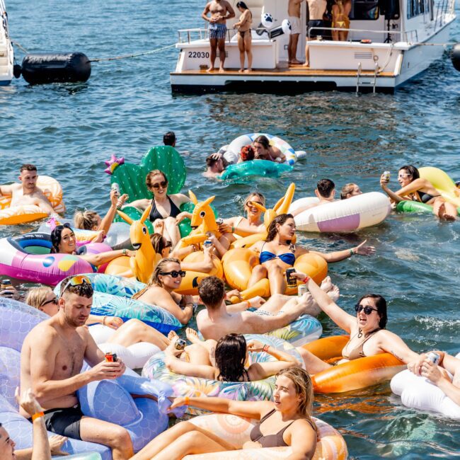 A lively group of people is enjoying a sunny day on colorful inflatable pool floats in the water near a yacht. The yacht in the background has more people onboard, some looking at the scene. The atmosphere is festive and relaxed.