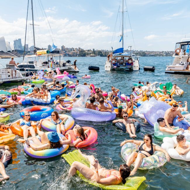 A lively scene of people on colorful inflatable floats in the water near several boats. The floats include various shapes like unicorns and donuts. The background shows a city skyline under a clear blue sky.