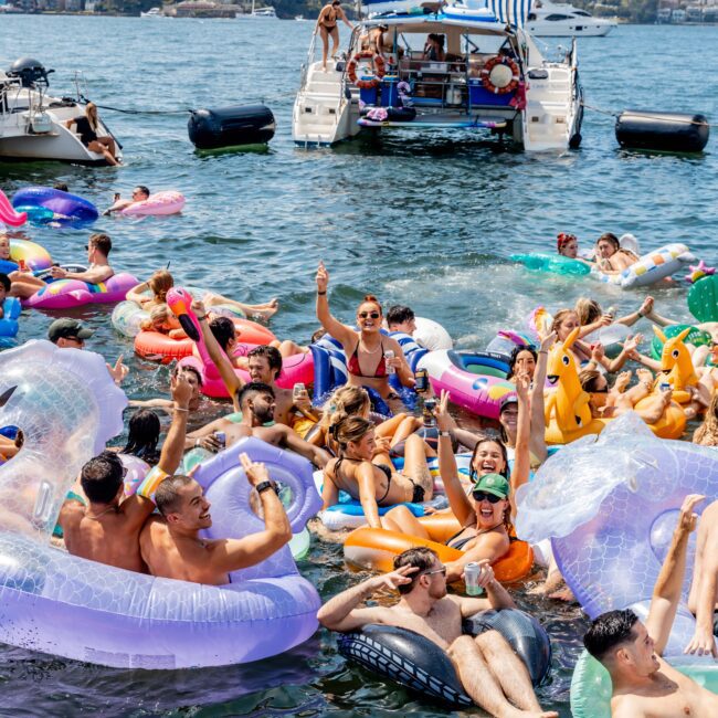 A lively scene with a crowd of people enjoying a sunny day on colorful pool floats in the water. A white boat with a blue-striped canopy is anchored nearby, and people on the boat and in the water are smiling and waving.