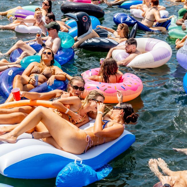 A crowded lake scene with people relaxing on inflatable floats of various shapes like flamingos, donuts, and whales. Some individuals hold drinks, wearing swimwear and sunglasses, enjoying a sunny day in the water.