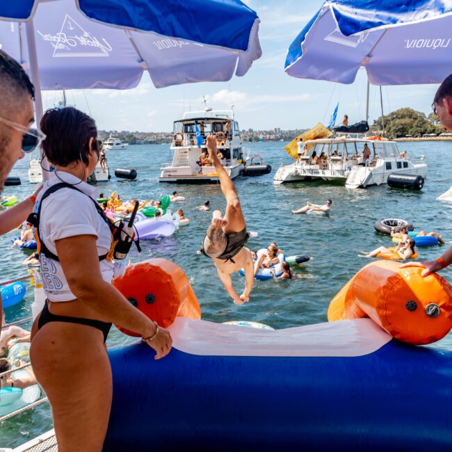 A group of people at a floating water park on a sunny day. One person is doing a backflip off an inflatable platform into the water. Others are on inflatables or watching from the platform. Boats and trees are in the background.