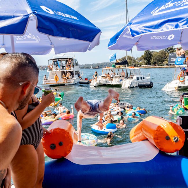 People enjoy a floating party with an inflatable slide on a sunny day. A person is mid-air off the slide, while others watch and relax in the water with floaties. Boats and umbrellas are visible in the background.