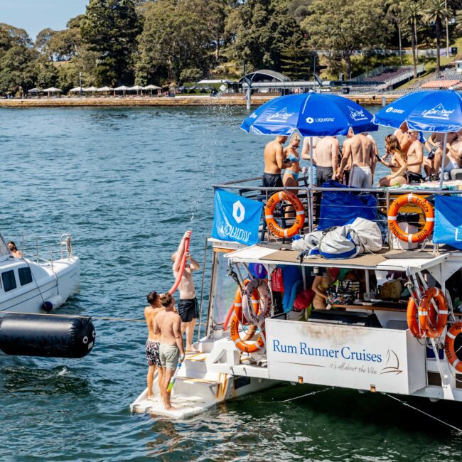 A crowded party boat, labeled "Rum Runner Cruises," is docked on a sunny day. People in swimwear are gathered on deck, with some entering the water. Nearby, a smaller boat is visible. Trees line the background.