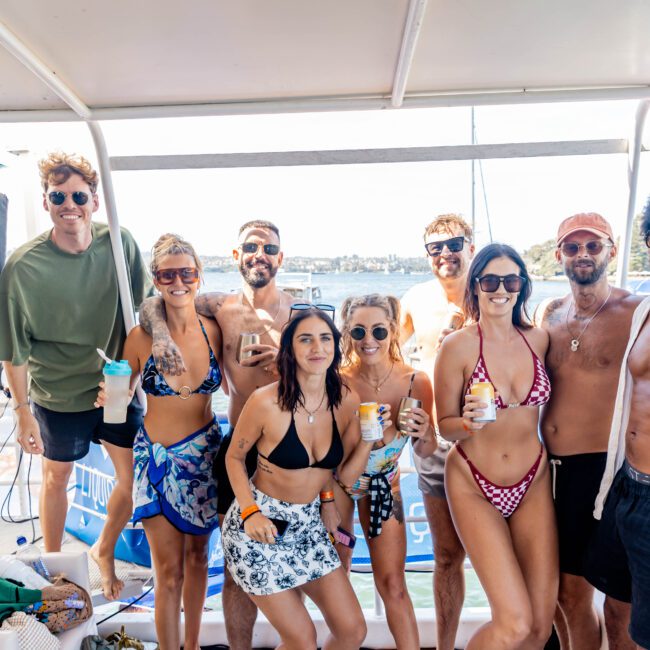 A group of nine people stands on a boat, smiling and holding drinks. They are dressed in swimwear. Sunlight is streaming in, and the water is visible in the background. Some wear sunglasses.