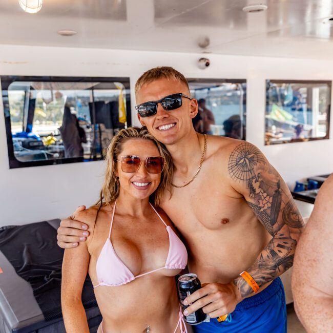 A man and woman in swimwear smile and pose together on a boat. The woman is wearing a pink bikini and sunglasses, while the man, with tattoos, wears blue shorts and sunglasses. They’re both holding drinks with a relaxed and happy demeanor.