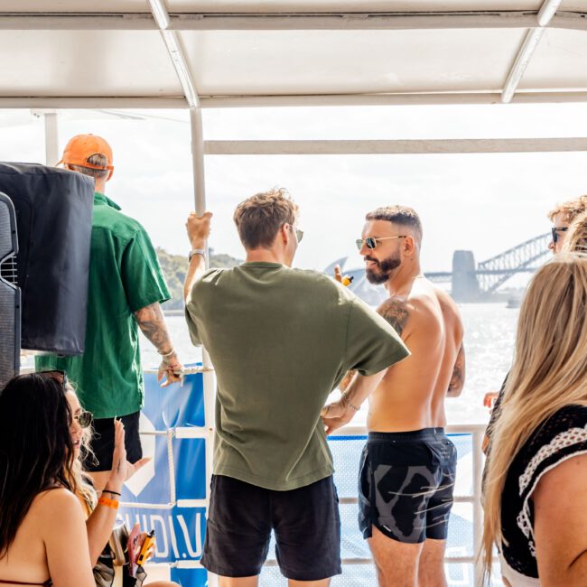 A group of people enjoying a boat party on a sunny day. Some are standing by the railing, while others are seated. The background features a bridge over a shimmering body of water. One woman in the foreground holds a drink.