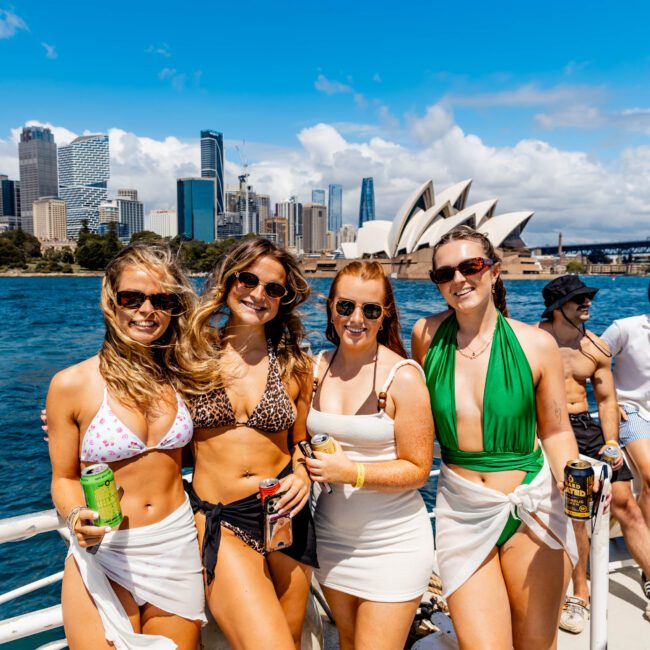 Four women in swimsuits and sarongs pose on a boat with the Sydney Opera House and city skyline in the background. The sky is blue with some clouds. They are holding drinks and smiling at the camera.