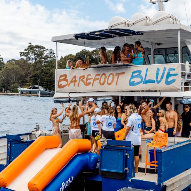 A group of people is having a party on a boat named "Barefoot Blue" by the water. They are mingling, some seated and some dancing. The boat features an inflatable slide. Trees and a shoreline can be seen in the background.