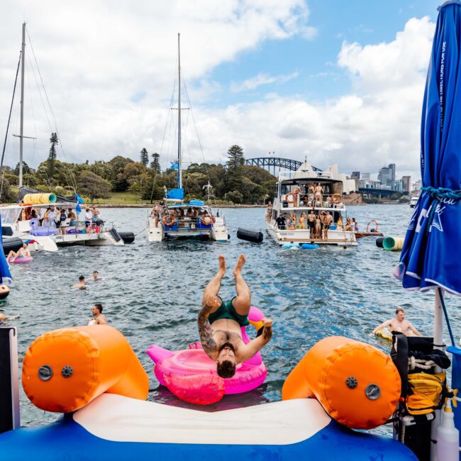 A man performs a backflip into the water from a pink inflatable in a lively bay setting. Boats and people float nearby. Blue umbrellas are in the foreground, and a bridge and city skyline are visible in the background.