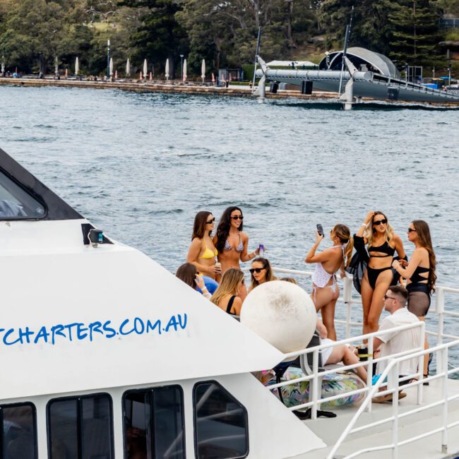 A group of people are socializing on a boat near the shore. They are wearing swimsuits and enjoying a sunny day. The water is calm, and the background shows trees and a dock. The boat has visible markings of a charter service.