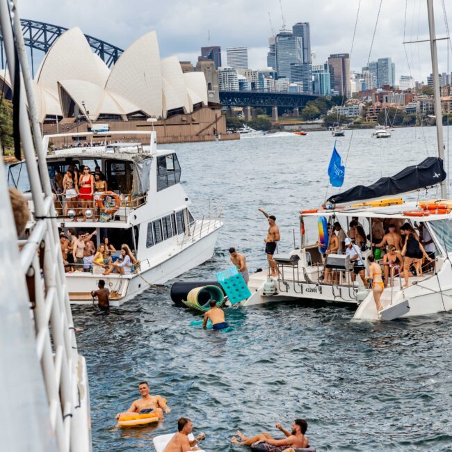 People are swimming and relaxing on inflatable floats near several boats on a sunny day, with the Sydney Opera House and Harbor Bridge in the background. The scene is lively, with individuals enjoying the water and socializing.