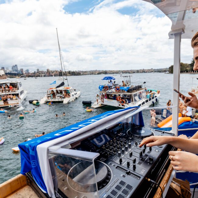 A woman wearing headphones is DJing on a boat deck surrounded by water. People are swimming and relaxing on nearby boats. The Sydney Opera House and Harbour Bridge are visible in the background under a partly cloudy sky.