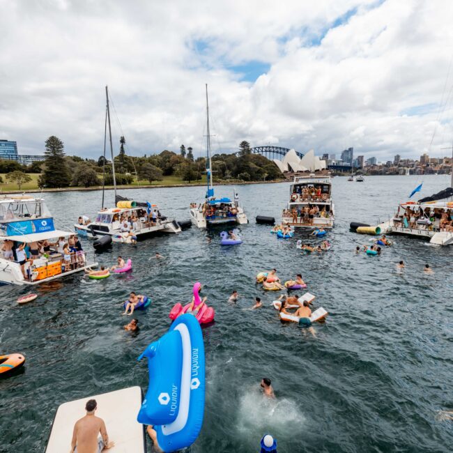 A group of boats anchored on a body of water with people swimming and floating on inflatables. The Sydney Opera House is visible in the background, along with city buildings and trees. The sky is partly cloudy.