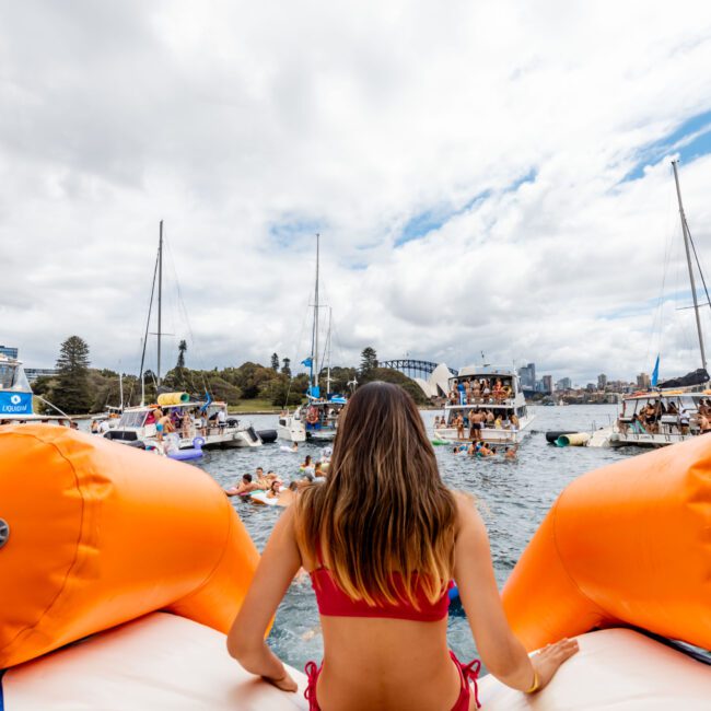 A person in a red swimsuit sits on an inflatable orange water toy, overlooking a lively water scene. Multiple boats and people are gathered in the water, with a cityscape and the edge of a bridge visible in the background.