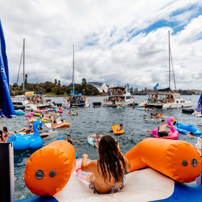 A vibrant gathering of people on colorful inflatable floats in a sunny harbor, surrounded by various boats. Blue and white umbrellas are visible on the boat, and the sky is partly cloudy.