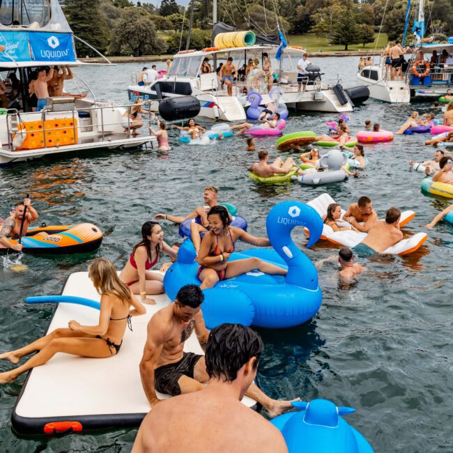 A lively scene on the water with numerous people on inflatable rafts and floats. Boats are anchored nearby, and people are swimming and socializing. Trees and a cloudy sky serve as a backdrop, creating a festive atmosphere.