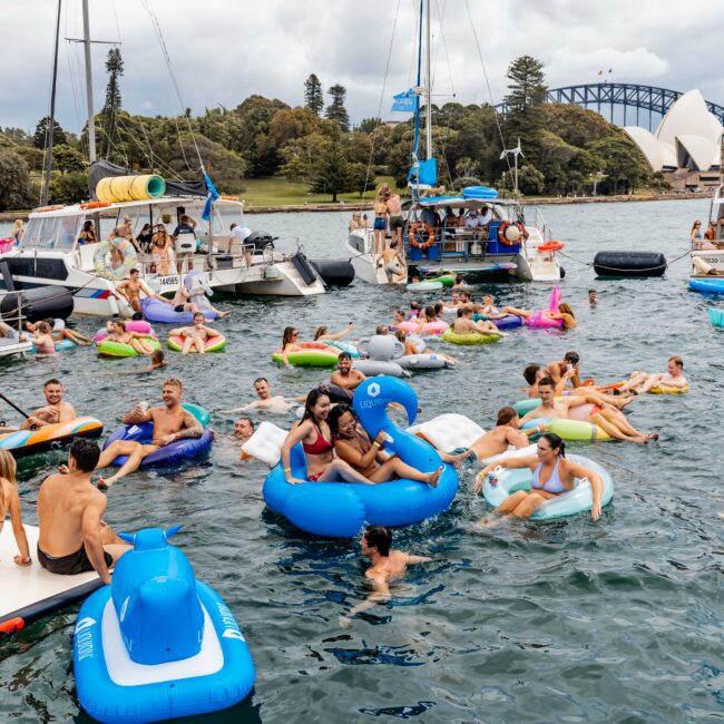 A lively scene of people enjoying a sunny day on the water, with several floating platforms and inflatables. In the background are boats with partygoers, lush trees, and Sydney's Opera House and Harbour Bridge in view.