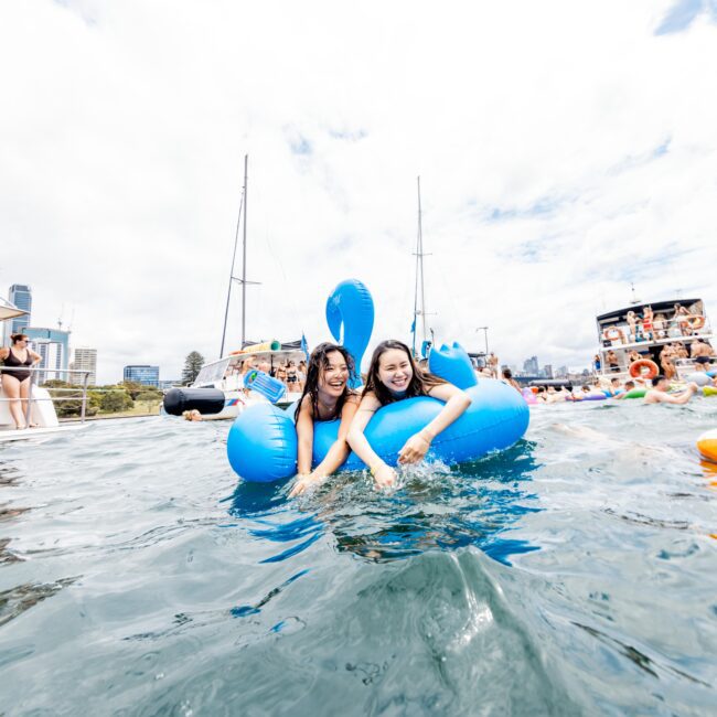 Two people smile and hug on a blue inflatable toy in the water. Boats with more people are in the background, under a partly cloudy sky. The scene suggests a fun, social event on the water.