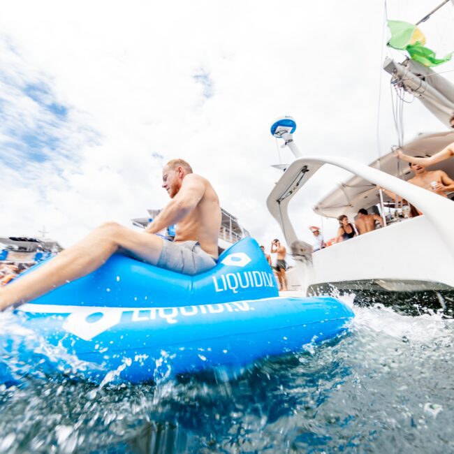 A man slides down a blue inflatable positioned at the side of a boat, splashing into the water. Others on the boat watch the action under a cloudy sky.