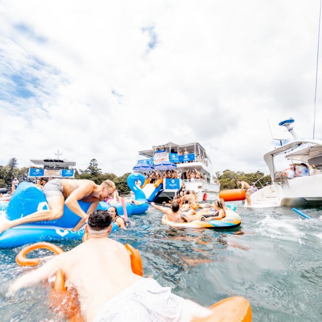 A group of people enjoying a sunny day on the water, lounging on colorful inflatable floats. A large boat with more people is in the background, and the sky is partly cloudy. The scene is lively and bright with a summer vibe.