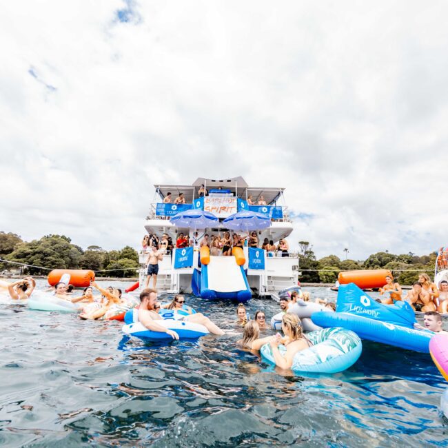 A lively scene on a lake with people enjoying a party on a boat. Many are in swimsuits, lounging on colorful floaties in the water. A large boat with banners and people is in the background, surrounded by trees and cloudy skies.