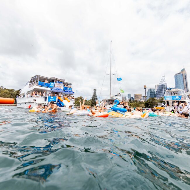 A large group of people on inflatables in the water near a boat. The boat is crowded with people, and city buildings are visible in the background under a cloudy sky.