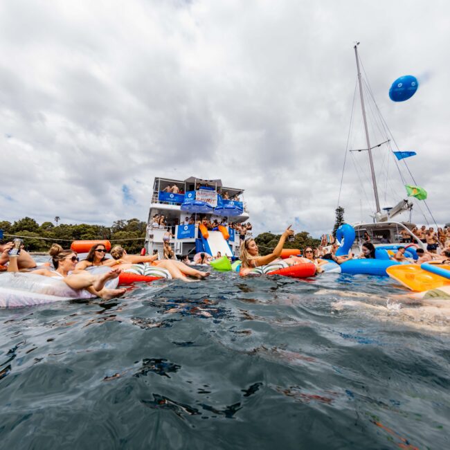 A lively scene on the water with people on inflatable floaties enjoying a party. The background features a large, multi-deck yacht adorned with banners and a nearby sailboat. The sky is cloudy, and a large blue beach ball is in the air.