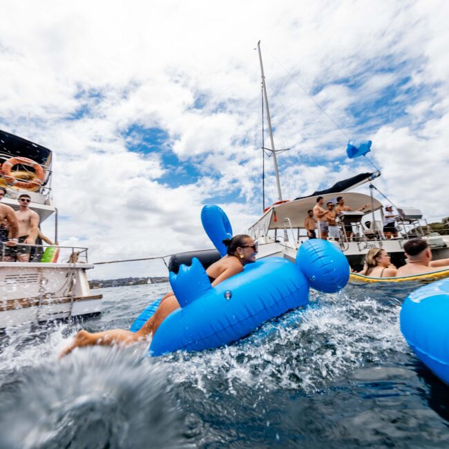 People are having fun on yachts, with some in the water on inflatable floats. One person is splashing water while riding a blue inflatable animal. The sky is partly cloudy and there are others on a nearby boat enjoying the scene.