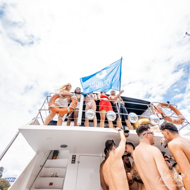 A group of people are partying on a yacht, with several individuals on the upper deck holding a blue flag. Others on the lower deck are shirtless, enjoying the sunny day. Orange life rings are attached to the railing. The sky is partly cloudy.