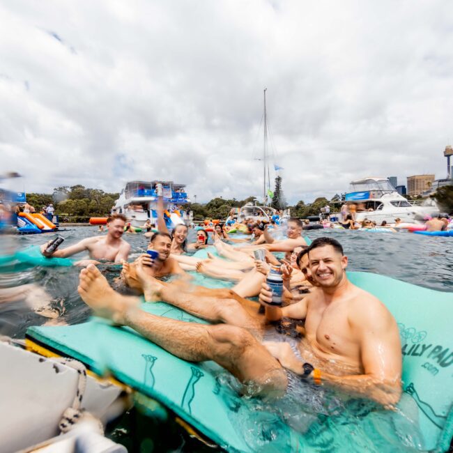 A group of people are relaxing on a large floating mat in the water, holding drinks and smiling. Boats and inflatables surround them on a sunny day. Buildings and trees are visible in the background under a partly cloudy sky.