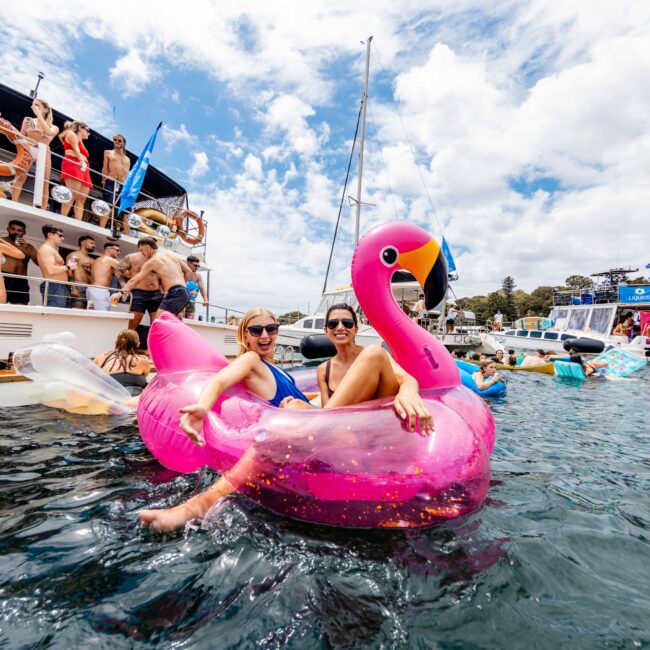 People are celebrating on a yacht. In the foreground, two people are sitting on a pink flamingo float in the water. Others are gathered on the boat, and several yachts and inflatables are seen in the background under a partly cloudy sky.