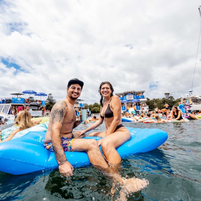 A man and woman smiling while sitting on a blue inflatable float in the water. They are surrounded by other people on floats and nearby boats. The background shows a cloudy sky and distant trees.