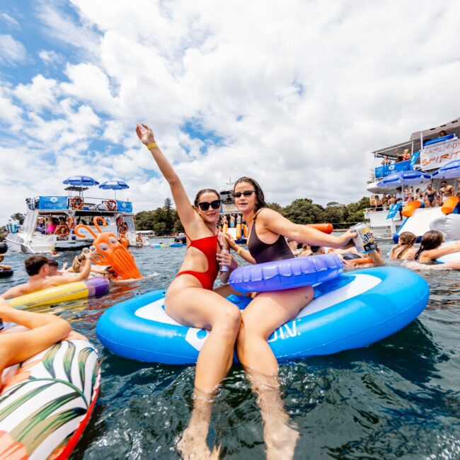 Two people sit on a blue inflatable float, smiling and posing for the camera. They are surrounded by others enjoying a sunny day on the water, and party boats are visible in the background. The scene is lively and festive.