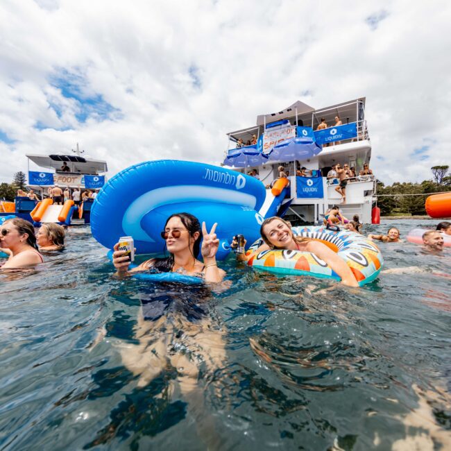 People are enjoying a sunny day on a lake, floating on inflatable tubes near a yacht. Some are holding drinks and smiling. The yacht is decorated with banners, and the scene is lively and festive under a partly cloudy sky.