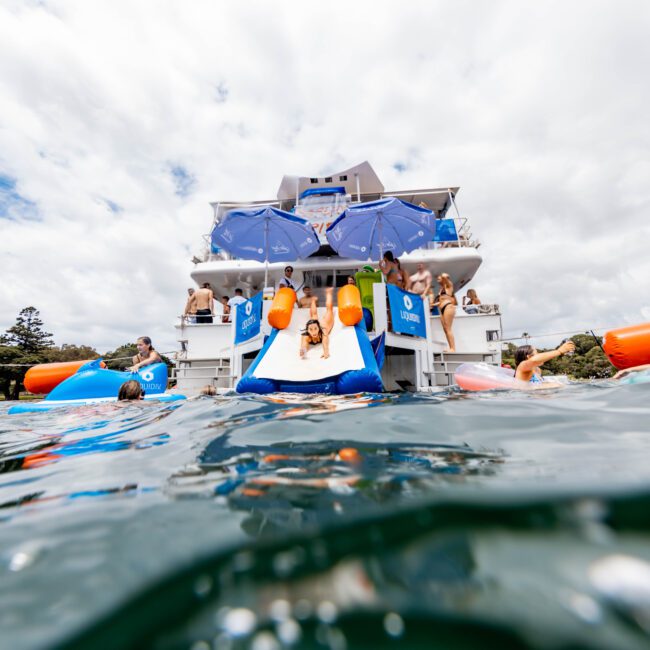 People enjoy a party on linked yachts. Some swim in the water, others sunbathe or use slides from the boats. An inflatable cooler with drinks floats nearby. The scene is lively and festive, set against a backdrop of trees and a cloudy sky.