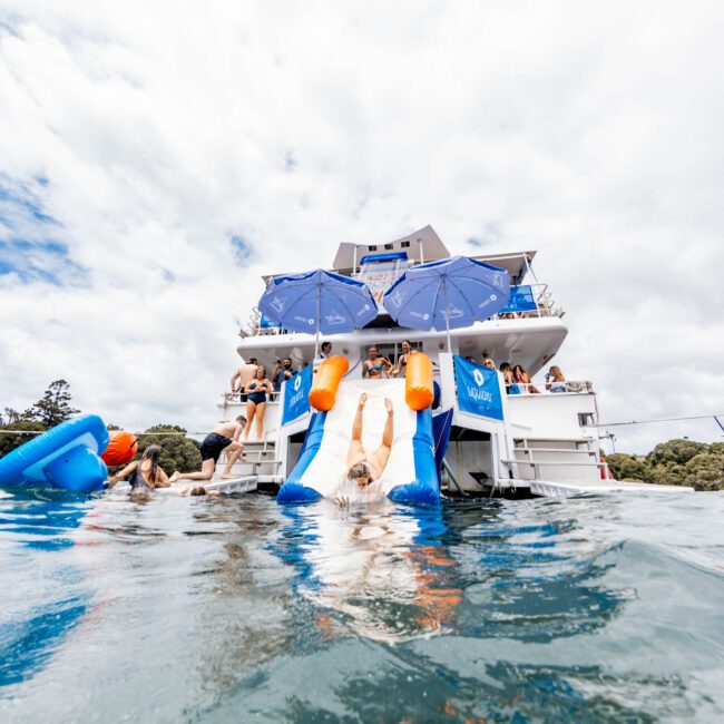 A person slides down a white and blue slide into the water from a yacht. The yacht has blue umbrellas and people are enjoying the day. The sky is partly cloudy.