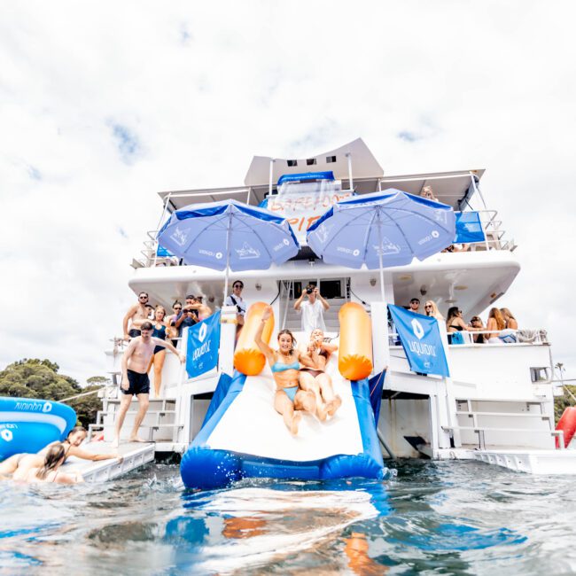 A group of people enjoy a bright day on a yacht, sliding down an inflatable water slide into the sea. The yacht features blue umbrellas, and others are standing and watching from the deck. The atmosphere is lively and festive.