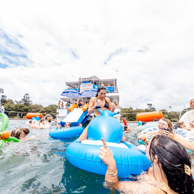 A lively scene on the water with several people enjoying themselves on inflatable floats near boats. The blue sky and fluffy clouds add to the cheerful atmosphere. People are relaxed and having fun in the sun.