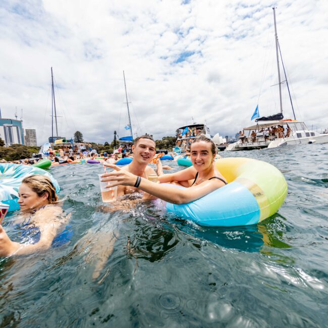 People enjoying a day in the water with inflatable tubes and drinks, near boats. The skyline of a city and cloudy skies are in the background. The scene is lively and festive.