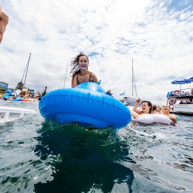 A woman joyfully rides a blue inflatable dolphin in the water, surrounded by others on similar floats. Several boats are anchored nearby under a partly cloudy sky.