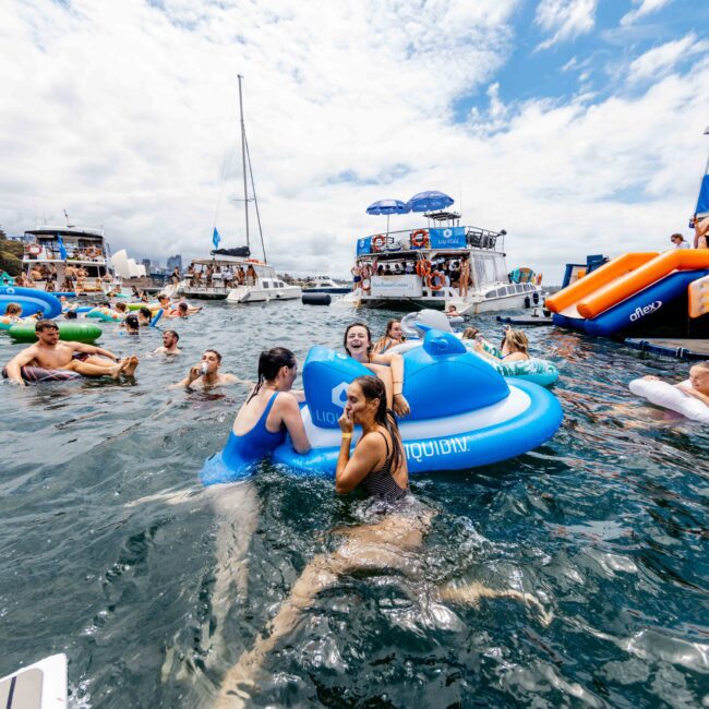 People are enjoying a sunny day in the water near several boats. Some are floating on inflatable toys, including a blue one and a white flamingo. The sky is partly cloudy, and the atmosphere is lively and fun.