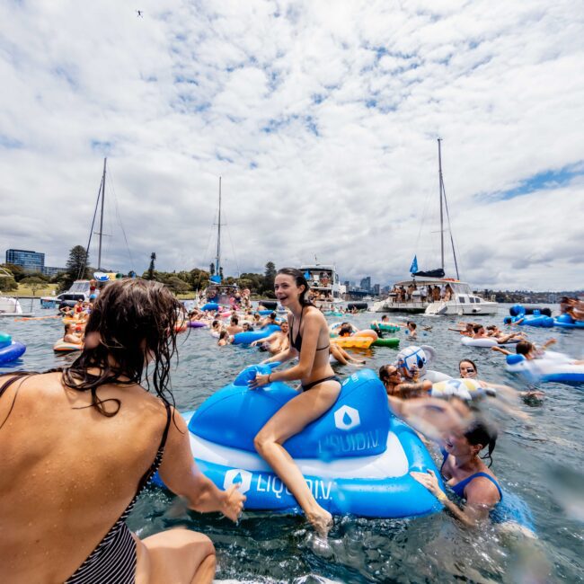 People are enjoying a sunny day on the water, surrounded by floating devices and boats. A woman in a striped swimsuit is in the foreground, while others are on blue inflatables. The sky is partly cloudy, and the water is lively with activity.