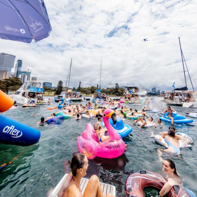 A lively gathering of people on colorful inflatable rafts and tubes in the water, with many wearing swimwear. Boats are visible in the background, and there's a partly cloudy sky above. Some participants are engaging with each other, enjoying the water.