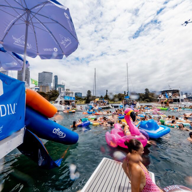 A lively yacht party with many people enjoying the water on inflatable floats of various shapes and colors. A large "Liquidn" banner is visible. Boats are anchored nearby under a partly cloudy sky with a small plane overhead.