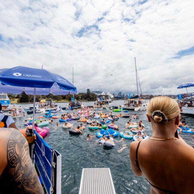 A crowd of people floating on inflatable pool toys in a body of water during a party. Boats with blue umbrellas are seen in the background, and two people stand on a platform in the foreground. The sky is cloudy, with a city skyline visible in the distance.