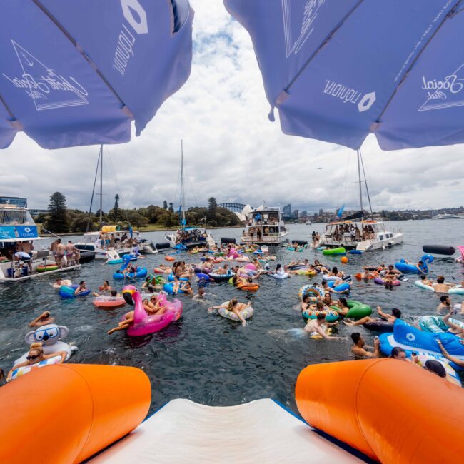 A lively outdoor scene with numerous people on colorful inflatables in the water, surrounded by boats. Two orange slides are in the foreground. Above are large white umbrellas with logos. Overcast sky and city buildings in the background.