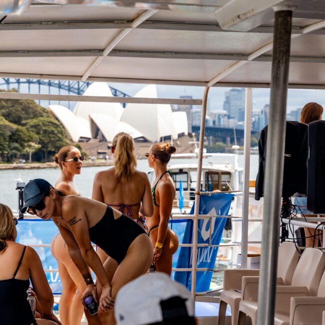 A group of people in swimsuits stand on a boat deck, with the Sydney Opera House and Harbour Bridge in the background. Some are posing and others are seated. A person in a cap is seen in the foreground, partially obscured.