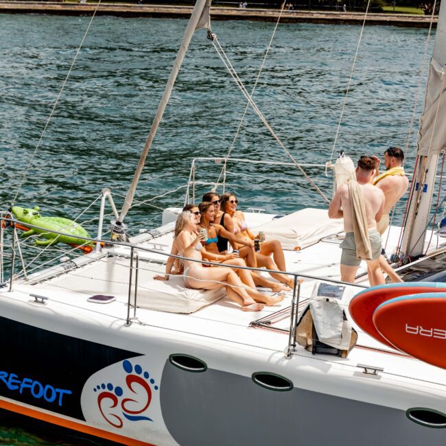 A group of people relax on the deck of a sailboat named "Barefoot" on the water. Some are seated, while one stands near a railing. A canoe is secured on the deck. The water is calm and another shoreline is visible in the background.