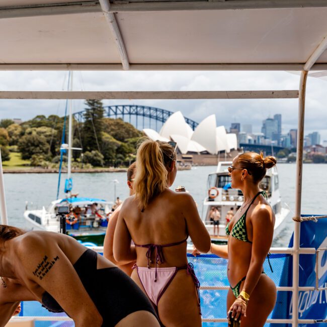 A group of people on a boat in swimsuits, with the Sydney Opera House and Harbour Bridge visible in the background. Some are chatting while others look at the view. The sky is cloudy.